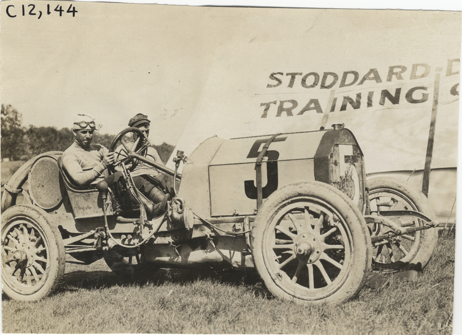 Driver and passenger in racecar in front of Stoddard-Dayton tent ...