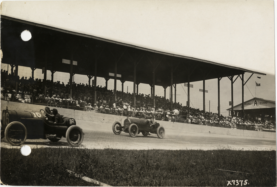 Mercer and Peugeot cars racing past grandstand, 1913 Indianapolis 500 automobile race