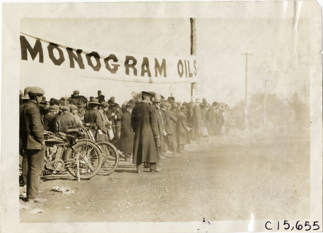 Spectators standing under Monogram oils banner, 1909 Vanderbilt Cup races