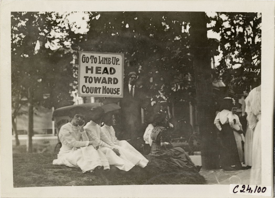 Women spectators sitting under sign, 1910 Glidden Tour